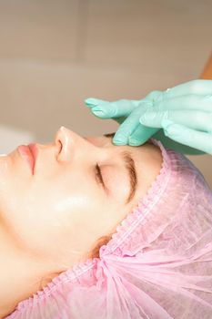 Cosmetologist with gloved hands applies a moisturizing mask with peeling cream on the female face. Facial cosmetology treatment. Procedures for facial care