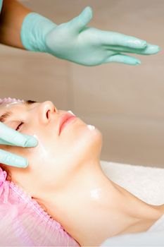 Cosmetologist with gloved hands applies a moisturizing mask with peeling cream on the female face. Facial cosmetology treatment. Procedures for facial care