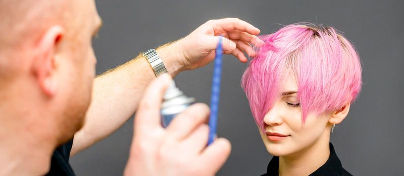 The hairdresser is using hair spray to fix the short pink hairstyle of the young caucasian woman in the hair salon