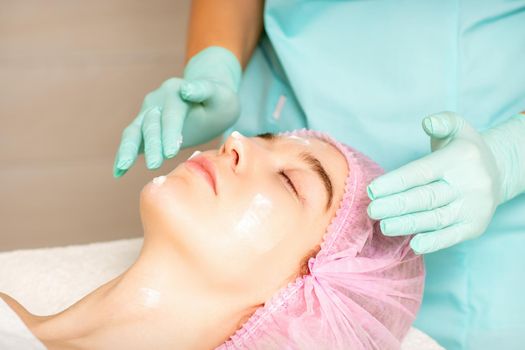 Cosmetologist with gloved hands applies a moisturizing mask with peeling cream on the female face. Facial cosmetology treatment. Procedures for facial care