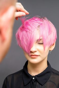 Hairdresser with hands is checking out and fixing the short pink hairstyle of the young white woman in a hair salon