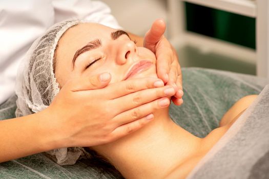 Beautiful caucasian young woman receiving a facial massage with closed eyes in spa salon, close up. Relaxing treatment concept