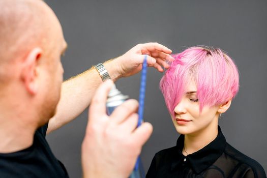 The hairdresser is using hair spray to fix the short pink hairstyle of the young caucasian woman in the hair salon