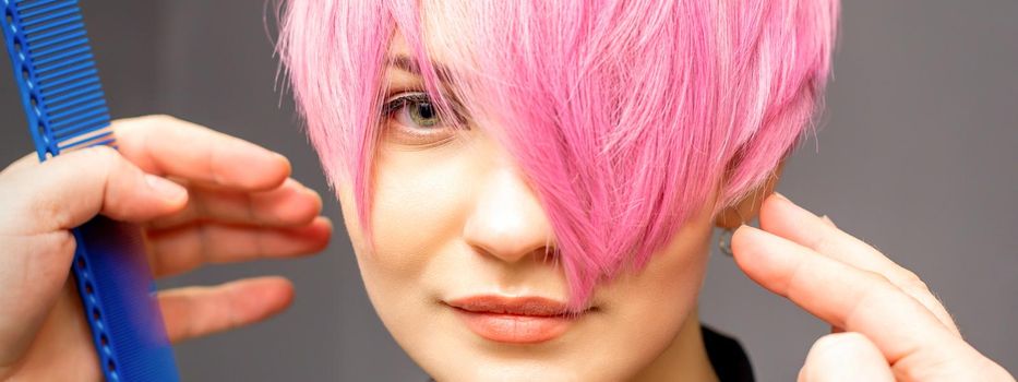 Hairdresser with hands and comb is checking out and fixing the short pink hairstyle of the young white woman in a hair salon