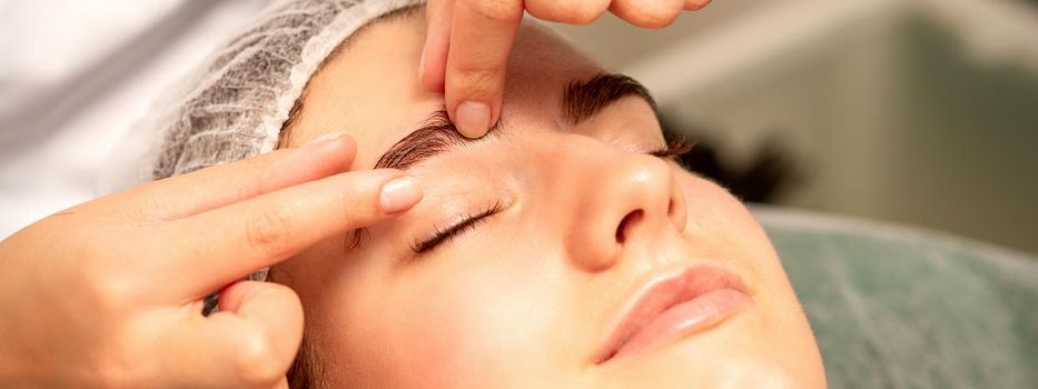 Eyebrow massage. Beautiful caucasian young white woman receiving an eye and eyebrow massage with closed eyes in a spa salon