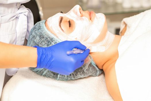 The beautician with brush applies a photochemical and glycolic peeling face mask to the female patient face in the beauty clinic