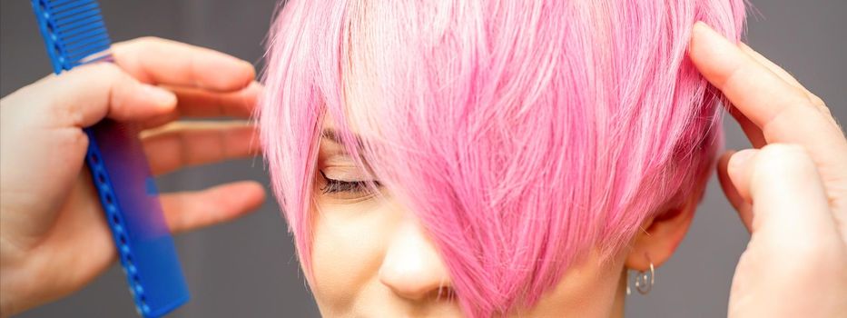 Hairdresser with hands and comb is checking out and fixing the short pink hairstyle of the young white woman in a hair salon