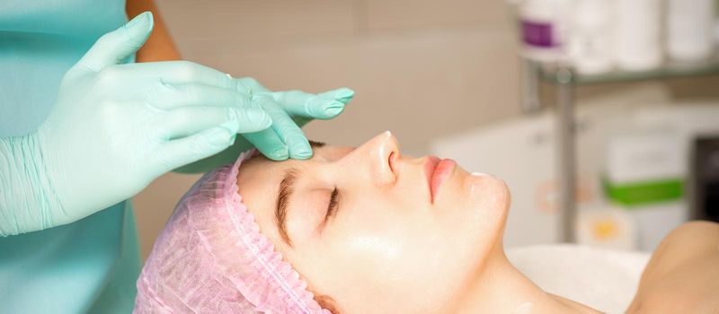 Cosmetologist with gloved hands applies a moisturizing mask with peeling cream on the female face. Facial cosmetology treatment. Procedures for facial care