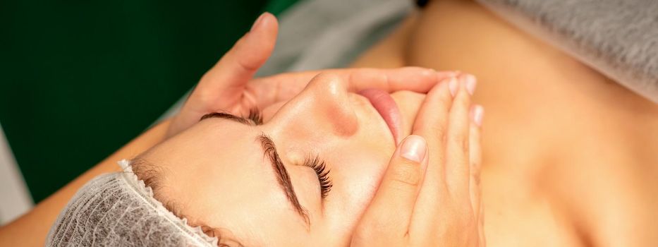 Beautiful caucasian young woman receiving a facial massage with closed eyes in spa salon, close up. Relaxing treatment concept