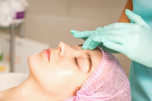 Cosmetologist with gloved hands applies a moisturizing mask with peeling cream on the female face. Facial cosmetology treatment. Procedures for facial care