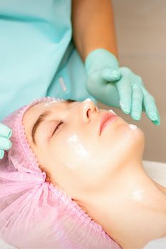 Cosmetologist with gloved hands applies a moisturizing mask with peeling cream on the female face. Facial cosmetology treatment. Procedures for facial care