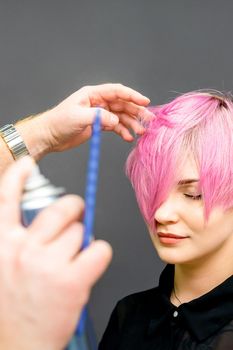 The hairdresser is using hair spray to fix the short pink hairstyle of the young caucasian woman in the hair salon