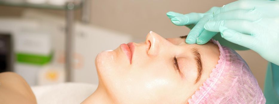 Cosmetologist with gloved hands applies a moisturizing mask with peeling cream on the female face. Facial cosmetology treatment. Procedures for facial care