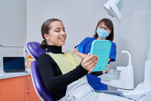 Happy young teenage female with mirror sitting in dentist chair smiling looking at healthy teeth, with dentist doctor in office. Dentistry, hygiene, treatment, dental health care concept