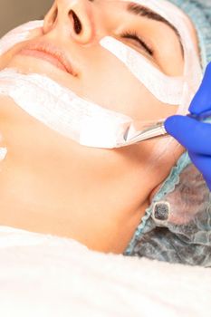 The beautician with brush applies a photochemical and glycolic peeling face mask to the female patient face in the beauty clinic