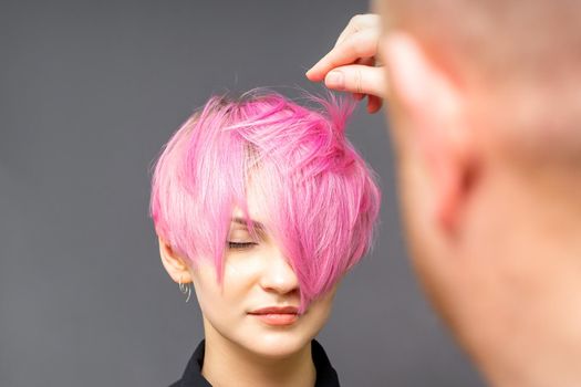 Hairdresser with hands is checking out and fixing the short pink hairstyle of the young white woman in a hair salon