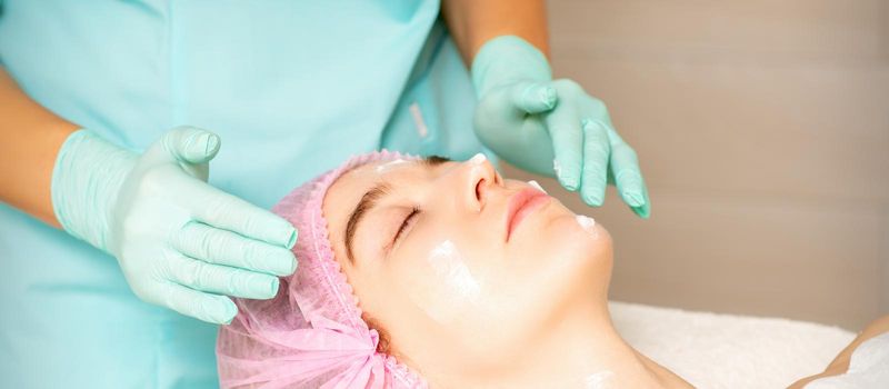 Cosmetologist with gloved hands applies a moisturizing mask with peeling cream on the female face. Facial cosmetology treatment. Procedures for facial care