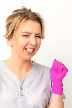 Happy caucasian woman doctor wearing pink gloves celebrates and raising fists on white background