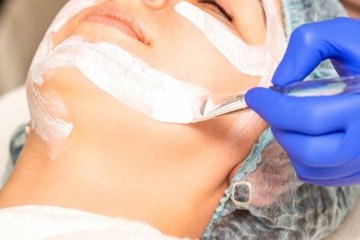 The beautician with brush applies a photochemical and glycolic peeling face mask to the female patient face in the beauty clinic