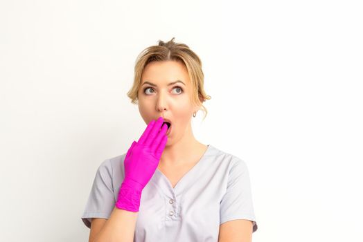 Portrait of a young female caucasian doctor or nurse is shocked covering her mouth with her pink gloved hands against a white background