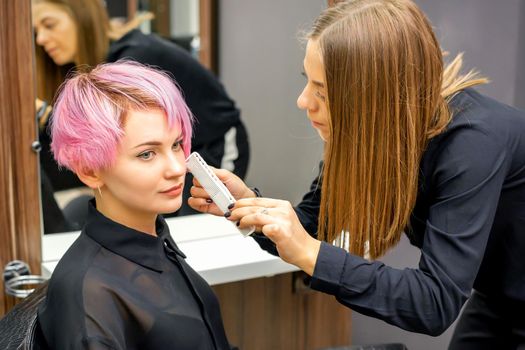 Hairdresser with comb is checking out and fixing the short pink hairstyle of the young white woman in a hair salon