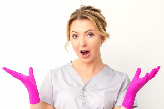 Body language. Young surprised Caucasian woman doctor wearing gloves gesturing with her hands, spread his arms having shocked expression opening her mouth against the white background