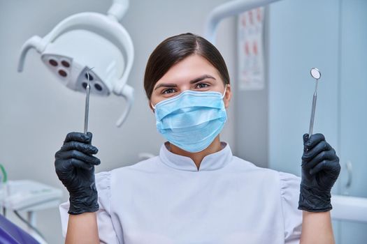 Headshot portrait of female dentist wearing facial mask with instruments for examining teeth. Doctor looking at camera, posing in dental office. Dentistry, medicine, treatment, dental health care