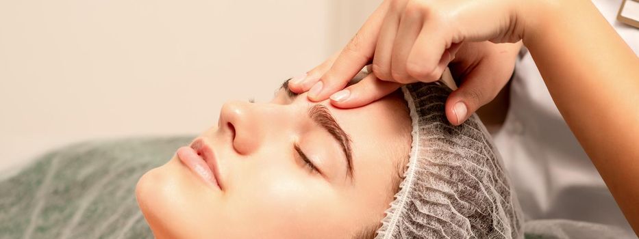 Beautiful caucasian young woman receiving a facial massage with closed eyes in spa salon, close up. Relaxing treatment concept