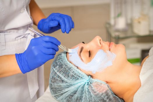 The beautician with brush applies a photochemical and glycolic peeling face mask to the female patient face in the beauty clinic