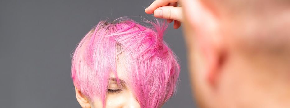 Hairdresser with hands is checking out and fixing the short pink hairstyle of the young white woman in a hair salon