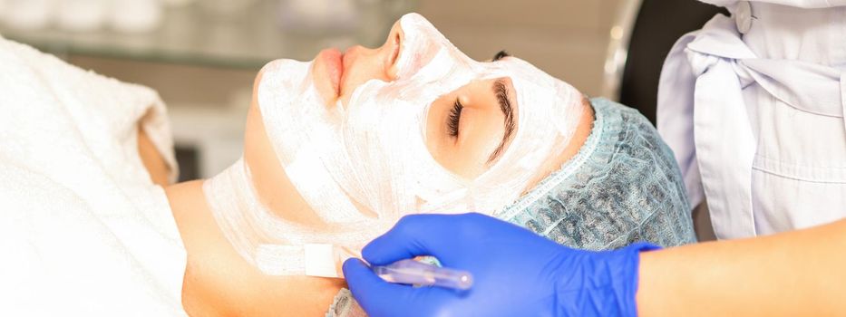 The beautician with brush applies a photochemical and glycolic peeling face mask to the female patient face in the beauty clinic