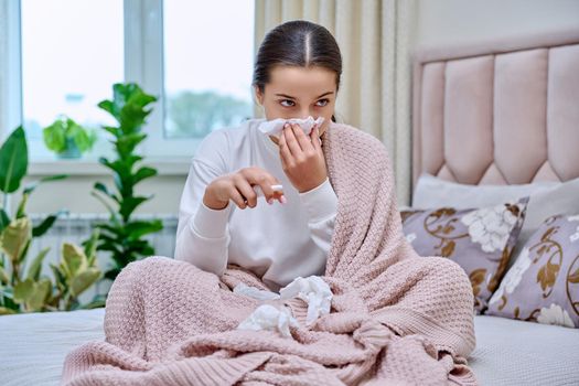 Young teenage female sitting in bed with runny nose medicine spray drops, sneezing into tissues, burying her nose. Autumn winter, rhinitis runny nose stuffy nose, treatment, flu season