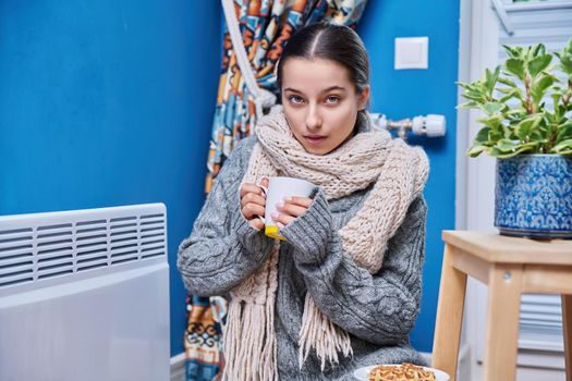 Cold autumn winter season, lifestyle, teenage female in warm sweater scarf warming near electric heating radiator, drinking hot drink with cup. Heating, warmth, equipment, life in cold season