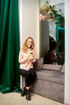 Beautiful caucasian young smiling woman with long wavy blonde hair holding and drinking a latte from a glass cup, sitting on a sofa