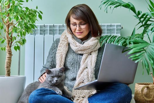 Middle-aged woman in warm clothes with cat using laptop. Female warming at home near heating radiator, with pet in her arms. Cold season winter autumn, leisure lifestyle work freelance, people animals