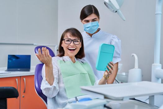 Happy middle aged woman together with dentist, patient sitting in dental chair looking at mirror. Prosthetics, treatment, implantation, dental teeth health, beauty care concept