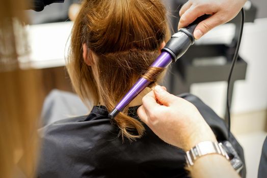 The hairstylist makes curls hairstyle of long brown hair with the curling iron in hairdresser salon, close up