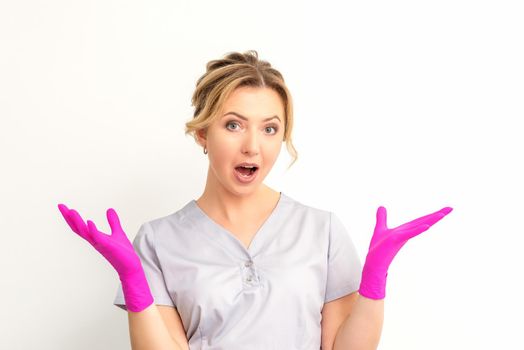 Body language. Young surprised Caucasian woman doctor wearing gloves gesturing with her hands, spread his arms having shocked expression opening her mouth against the white background