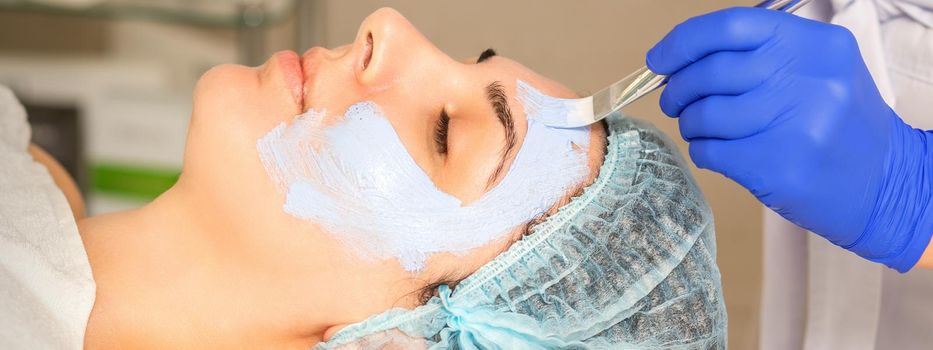 The beautician with brush applies a photochemical and glycolic peeling face mask to the female patient face in the beauty clinic