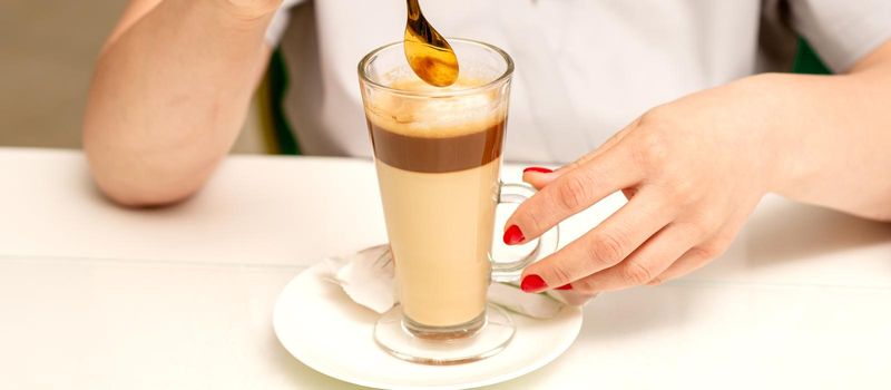 Woman with the latte. Glass mug of latte coffee on the white saucer with female hands holding teaspoon on the table in the cafeteria, close up
