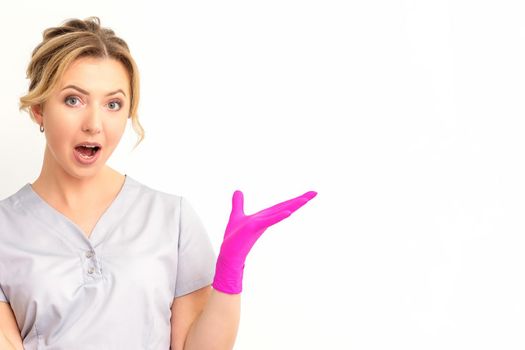 Body language. Young surprised Caucasian woman doctor wearing gloves gesturing with her hands, spread his arms having shocked expression opening her mouth against the white background