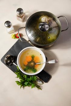 A bowl and a pot with fresh home made chiken broth with carrot and spices on black cutting board on white table, flat lay, top view.