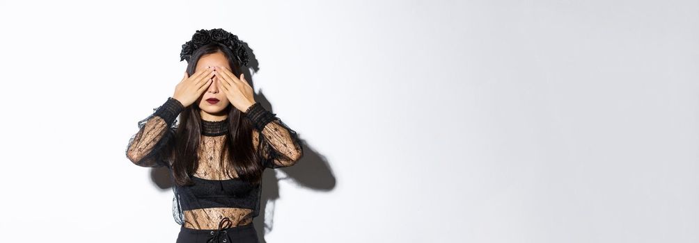 Image of woman wearing gothic wreath and black lace dress shut her eyes with hands, waiting for surprise on halloween, standing over white background with patient expression.