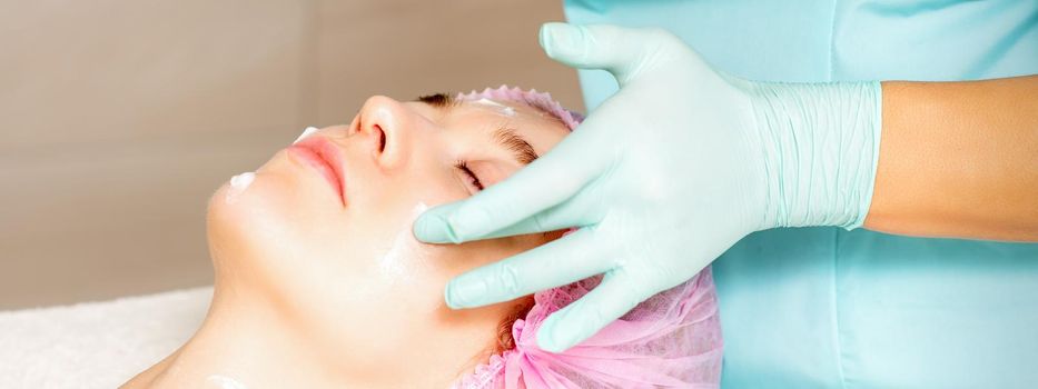 Cosmetologist with gloved hands applies a moisturizing mask with peeling cream on the female face. Facial cosmetology treatment. Procedures for facial care