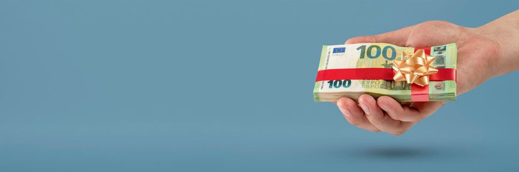 Gift of money. A man holds a stack of banknotes in his hands as a symbol or concept of a gift. 100 euro banknotes tied with a red ribbon with a bow on a blue background