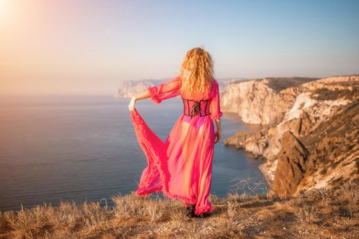 Side view a Young beautiful sensual woman in a red long dress posing on a volcanic rock high above the sea during sunset. Girl on the nature on blue sky background. Fashion photo