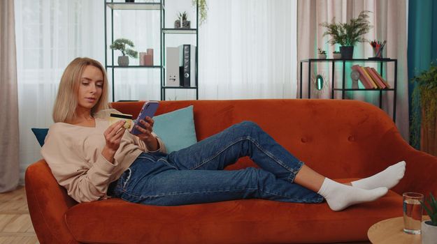 Happy shopaholic consumer young woman sitting at modern home apartment indoors making online order food delivery, payment with credit plastic bank card. Discount sale on internet. Adult girl on couch