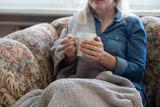 Close up of hands holding blue cup of tea or coffee. Model and cozy plaid is sitting, relax at home on sofa.