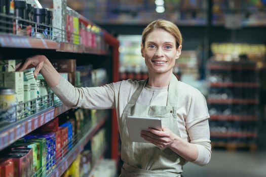 A young beautiful business woman, the owner of a store, a supermarket, stands in an apron near the shelves with products, holds a notebook in her hands, looks at the camera, smiles.