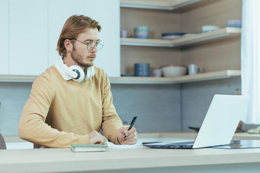 Young serious and focused man studying remotely at home, student in kitchen wearing glasses using laptop for distance learning taking notes on study material.